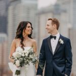 Elegant wedding couple holding hands in an urban rooftop setting with city skyscrapers in the background.