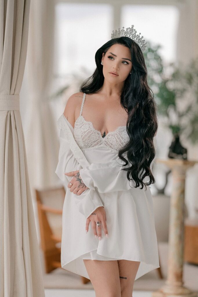 Luxury bride with long dark hair and crown, wearing elegant white satin robe, indoor portrait, high-end wedding styling, sophisticated bridal look.