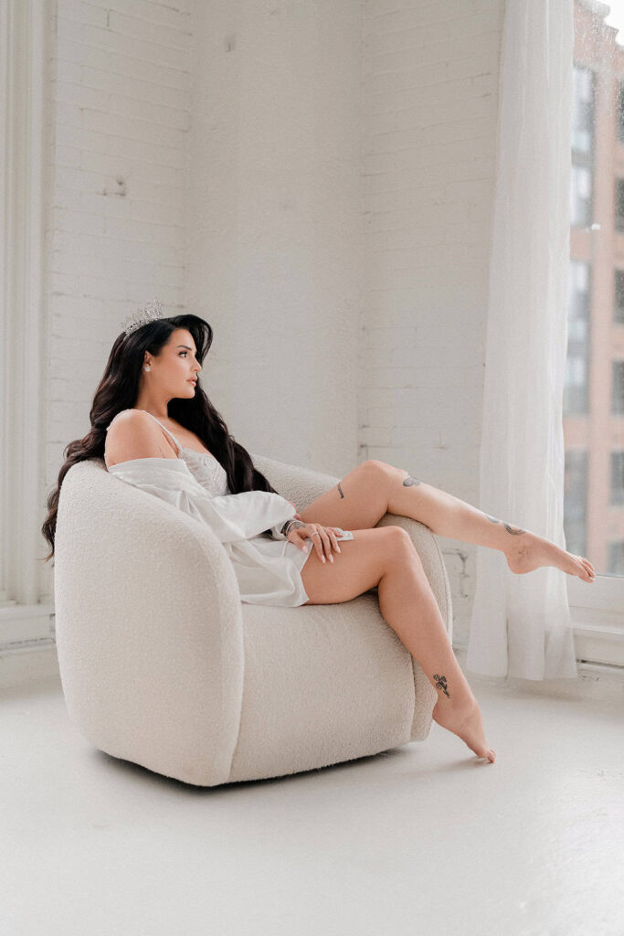 Elegant bride with dark hair and tattoos sitting on a white plush armchair in a bright, airy room with large windows and white brick walls.