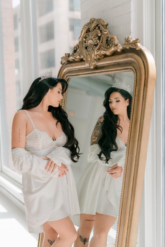 Elegant bride with dark hair and tattoos looking at her reflection in a vintage mirror, wearing a lace wedding slip and a tiara, in a high-end wedding venue.
