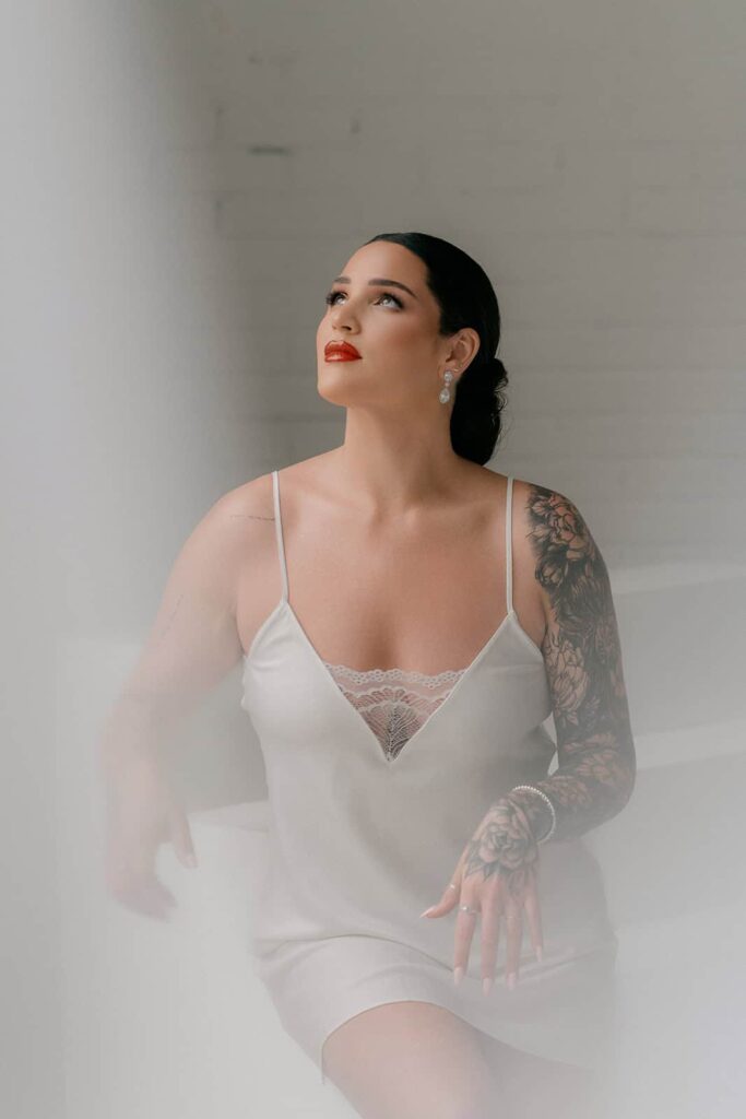 Elegant bride with dark hair and tattooed arms wearing a satin slip dress, posed in a minimalistic luxurious setting, showcasing sophisticated bridal style.