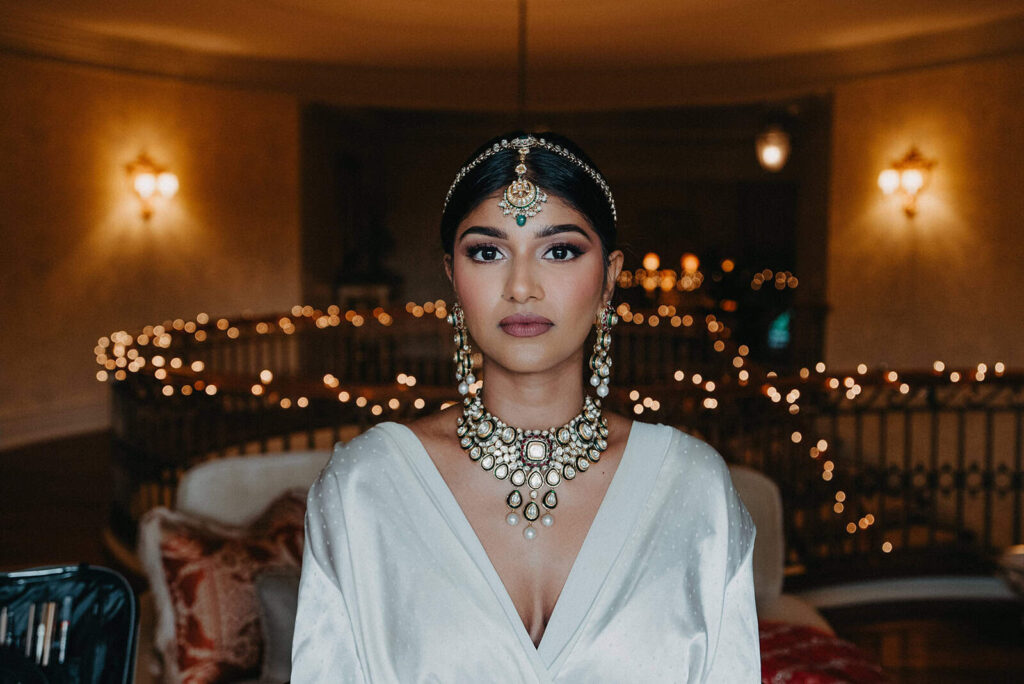 Elegant bride adorned with traditional jewelry at luxury wedding venue with ambient lighting and city skyline backdrop.