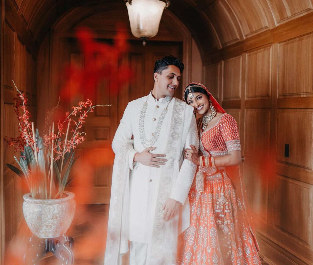 Elegant Indian bride and groom in traditional attire at luxurious wedding venue with warm wood-paneled walls and floral decor.