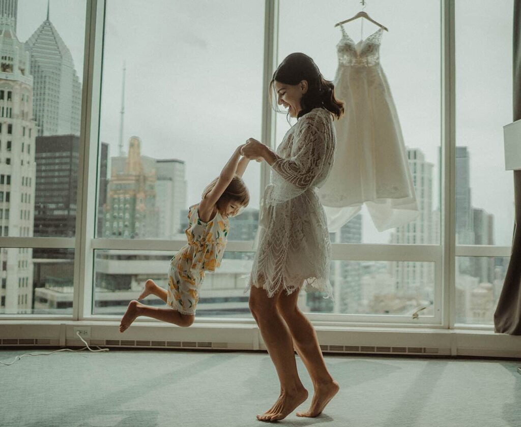 Elegant bride and young girl dancing in modern city high-rise apartment with wedding dress in background.