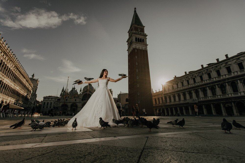 Elegant bride in wedding gown feeding pigeons at St. Mark's Square in Venice, Italy, during sunset.