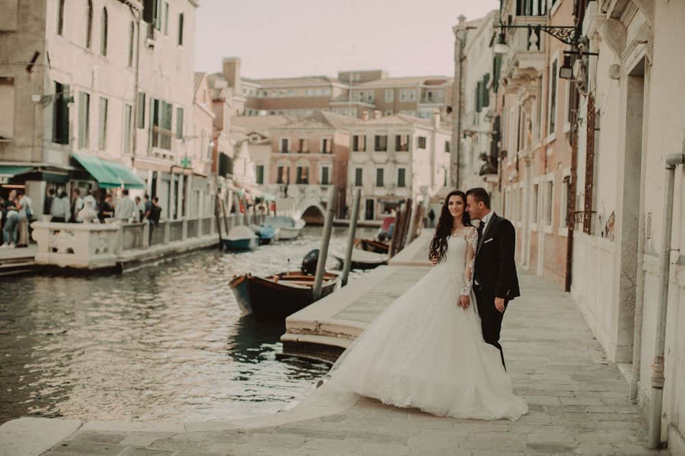 Elegant wedding couple in Venice canal with historic buildings and boats, showcasing luxury wedding photography.