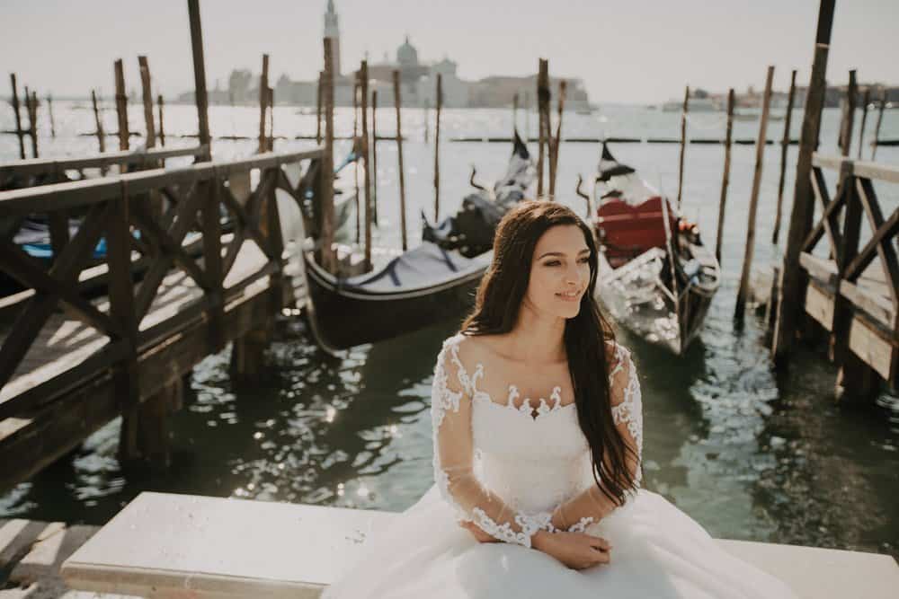 Elegant bride sitting by the water at a luxury wedding venue with docked boats, highlighting sophisticated wedding aesthetics.