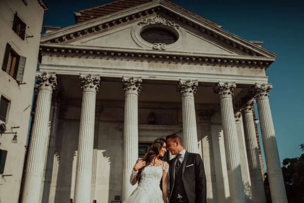 Elegant couple in wedding attire in front of a classical Greek-inspired wedding venue with tall white columns, perfect for luxury wedding photography.