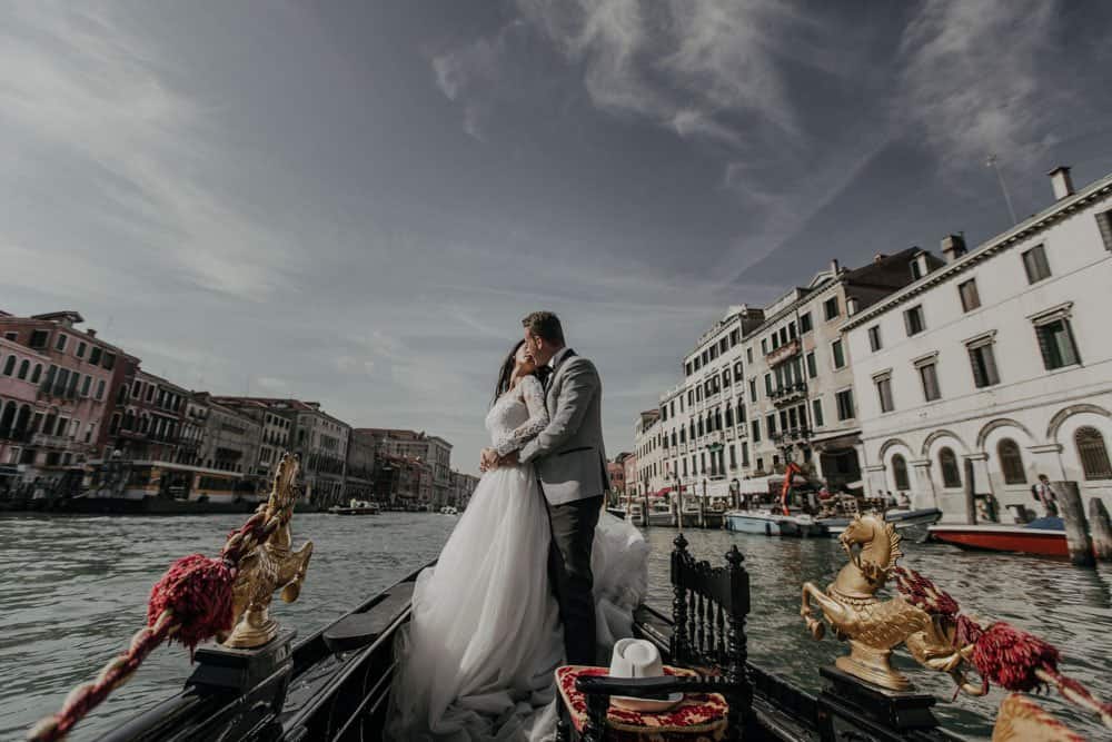 Elegant couple sharing a kiss on a gondola in Venice, surrounded by historic buildings and waterways, perfect for luxury wedding photography.
