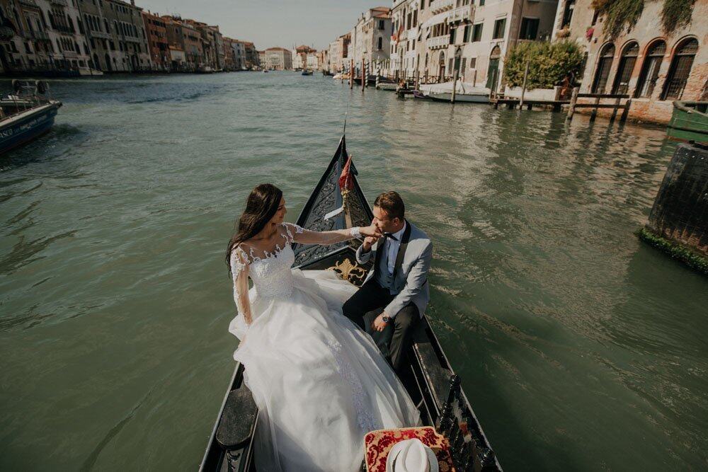 Elegant wedding couple on a gondola in Venice, with historic buildings surrounding the canal, creating a romantic and luxurious setting.