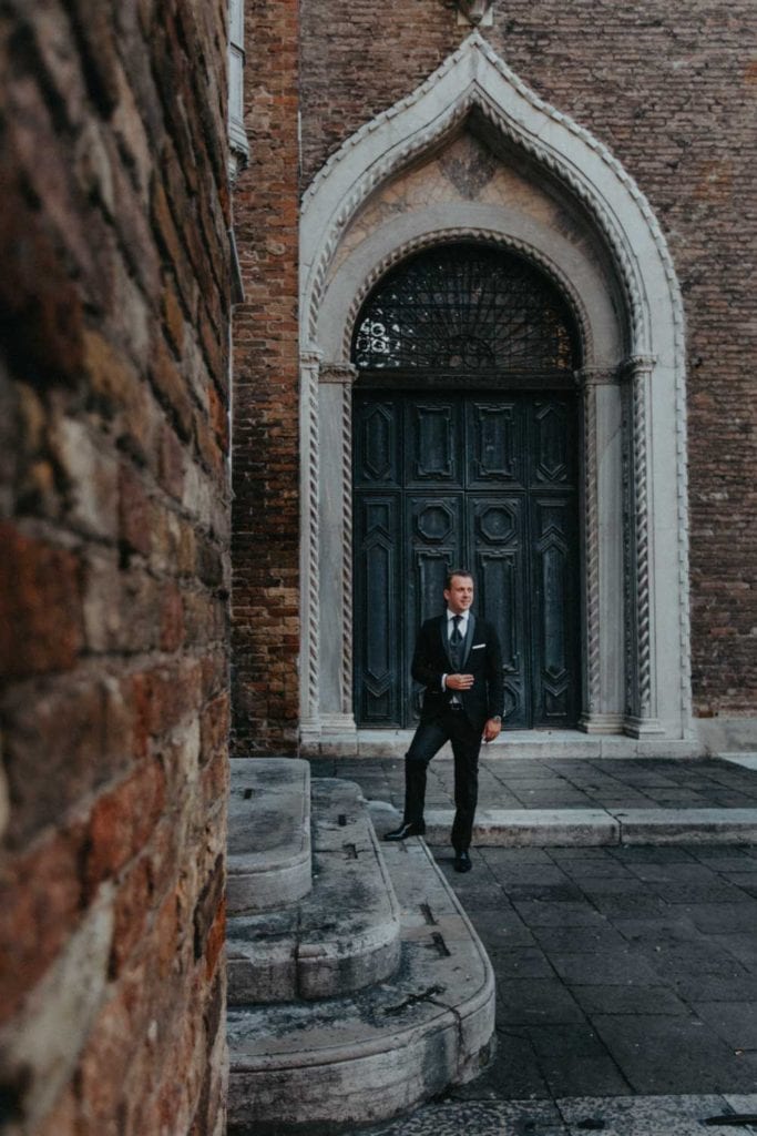 Elegant groom in tailored tuxedo stands outside historic church with ornate wooden door, perfect for luxury wedding photography.