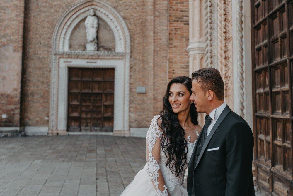 Elegant bride and groom in luxurious wedding attire outside historic venue looking at each other.