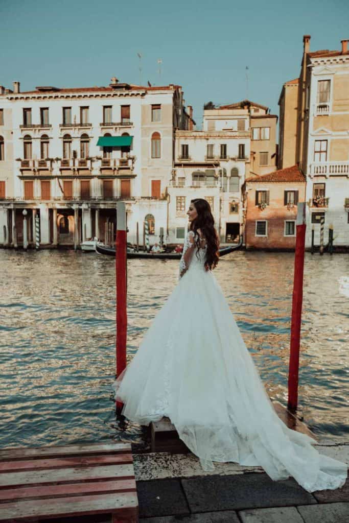 Venetian bride in wedding gown standing on dock by canal with historic buildings, Venice, Italy.