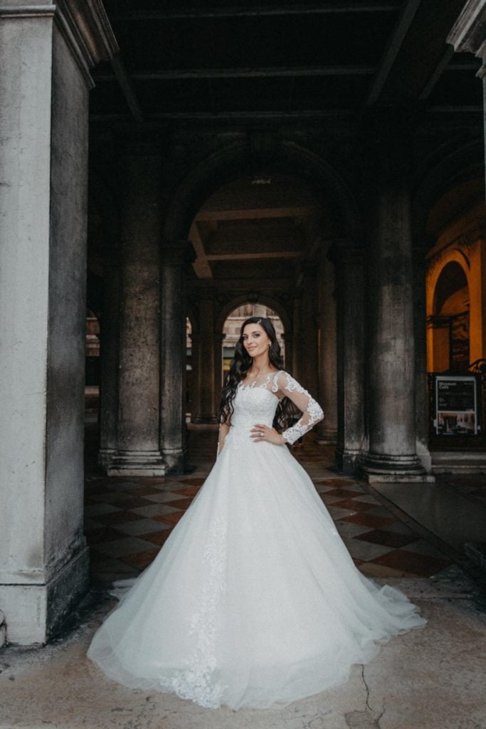 Elegant bride in a luxurious wedding gown standing in a historic high-end venue with grand arches and classical architecture.
