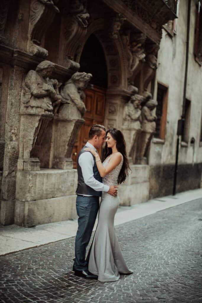 Elegant couple in wedding attire sharing a romantic moment outside a historic European building with ornate stone sculptures.