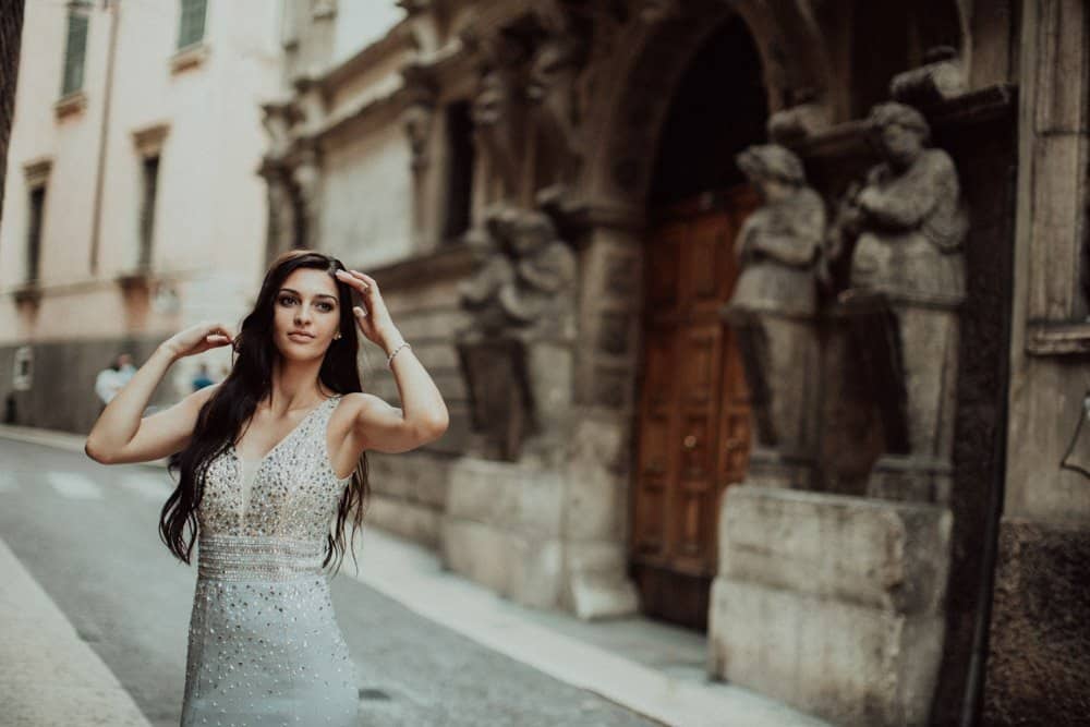 Elegant bride in luxury wedding dress standing in historic European street with ornate architecture and statues.