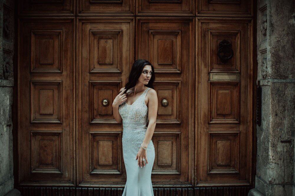 Elegant bride in a glamorous wedding gown standing against a historic wooden door at a high-end venue.