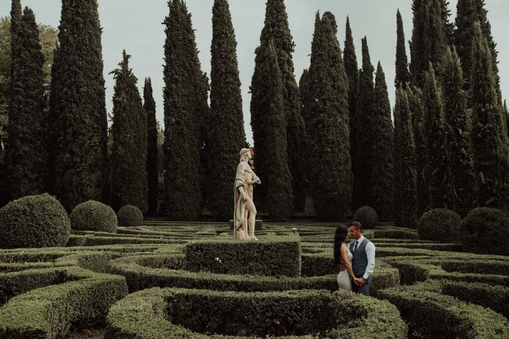 Luxurious wedding couple in elegant attire standing in a beautifully manicured garden with classical sculptures and tall cypress trees.