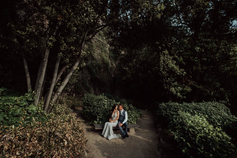 Elegant couple sitting on a park bench surrounded by lush greenery in a luxurious outdoor setting.