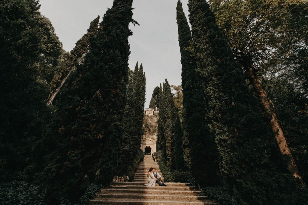 Majestic garden wedding scene with lush tall cypress trees and romantic couple sitting on stone steps, ideal for luxury wedding venues.