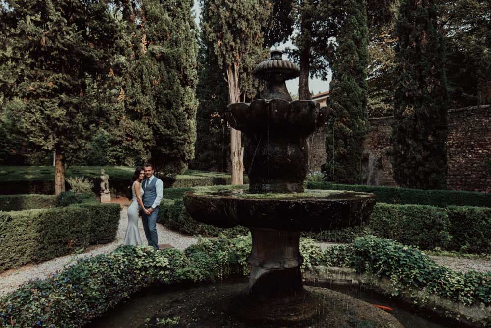 Elegant bride and groom in a lush garden wedding setting with classic stone fountain and manicured hedges.