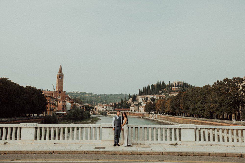 Beautiful couple in wedding attire on a picturesque bridge overlooking a charming European cityscape at sunset.