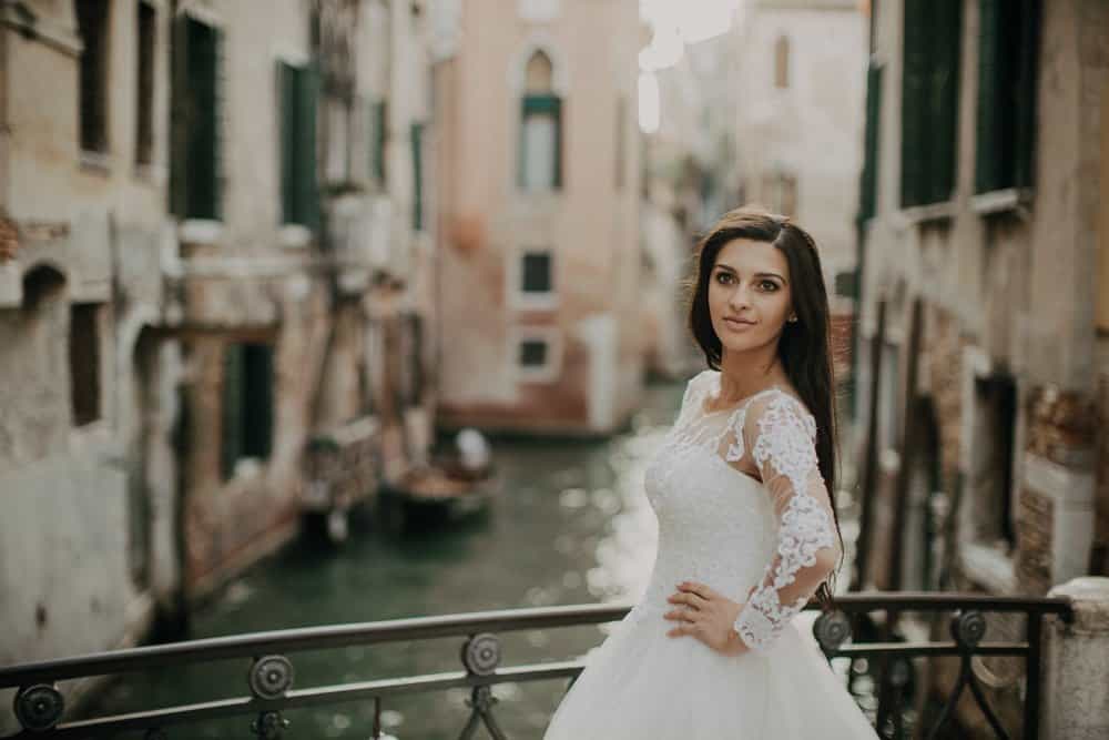Elegant bride in lace wedding dress on canal bridge in Venice, romantic high-end wedding venue.