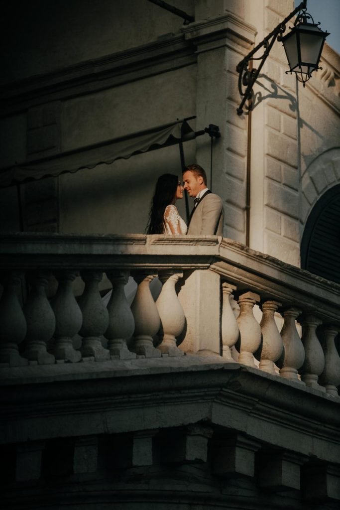 Elegant couple on a romantic balcony at a luxury wedding venue, with classical architecture and soft lighting enhancing the sophisticated atmosphere.