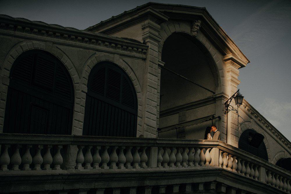 Elegant couple sharing a romantic moment on a luxurious balcony of a historic high-end venue at dusk, perfect for a wedding celebration.