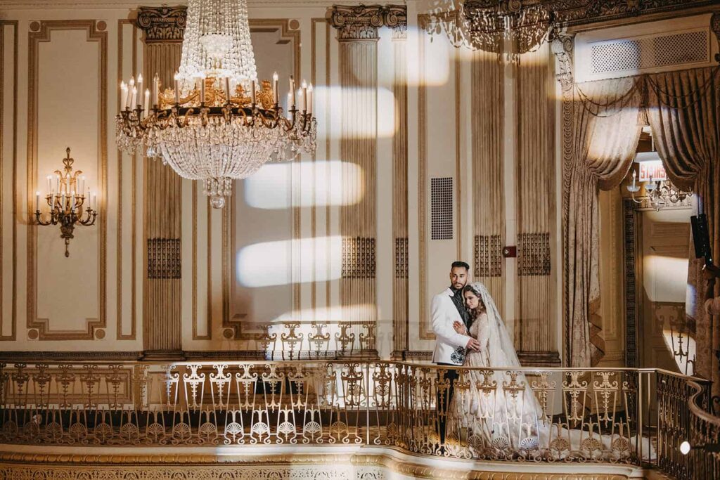 Elegant wedding couple in a luxurious historic venue with ornate gold details and grand chandeliers.