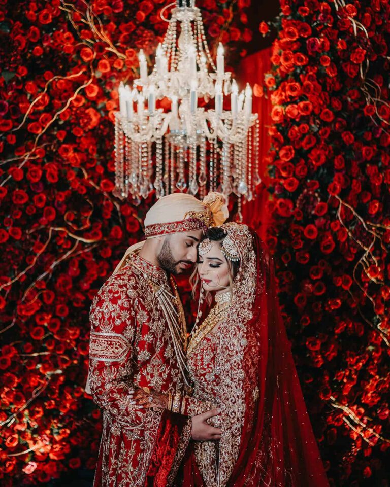 Beautiful wedding couple in traditional red Indian attire under a crystal chandelier surrounded by red roses.