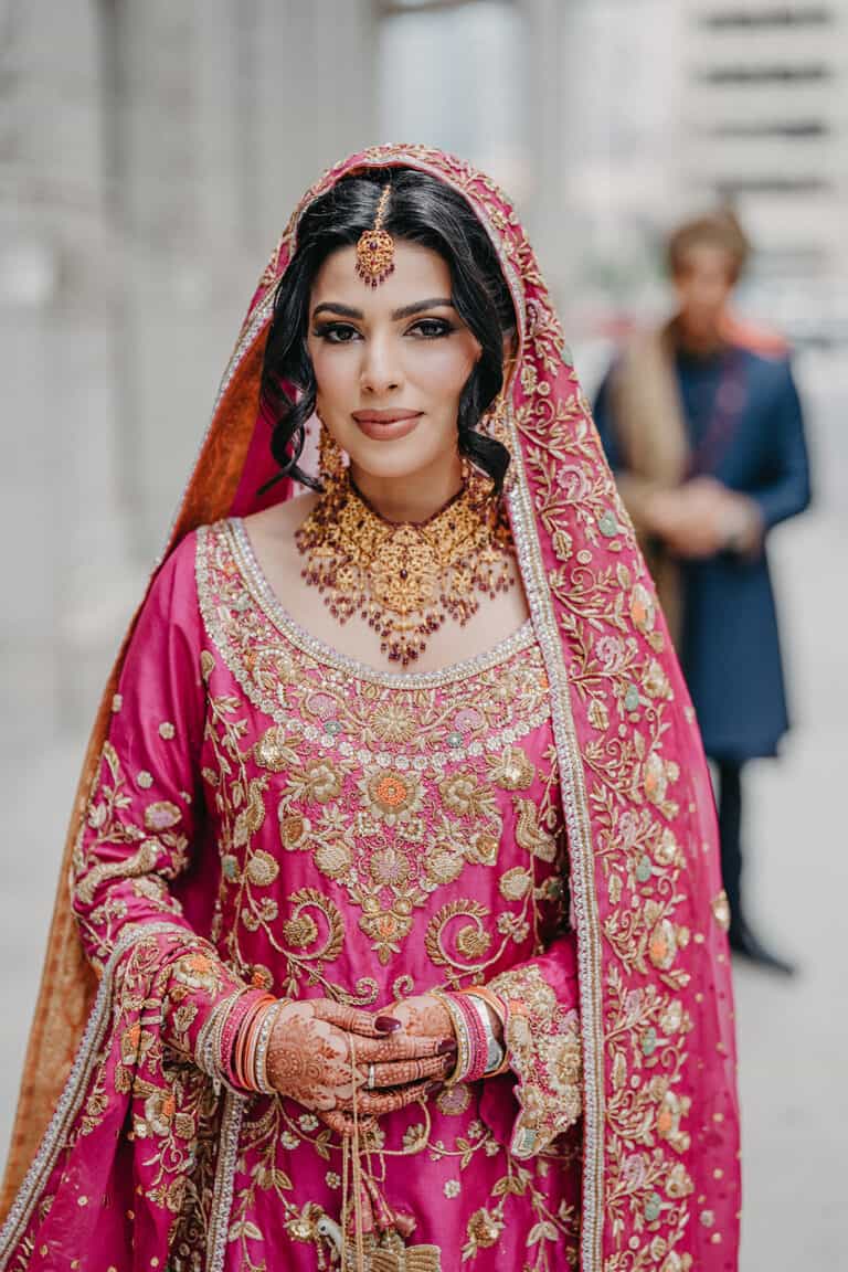 Stunning Muslim bride dressed in intricate pink and gold traditional wedding attire, adorned with jewelry, standing outdoors with cityscape background.
