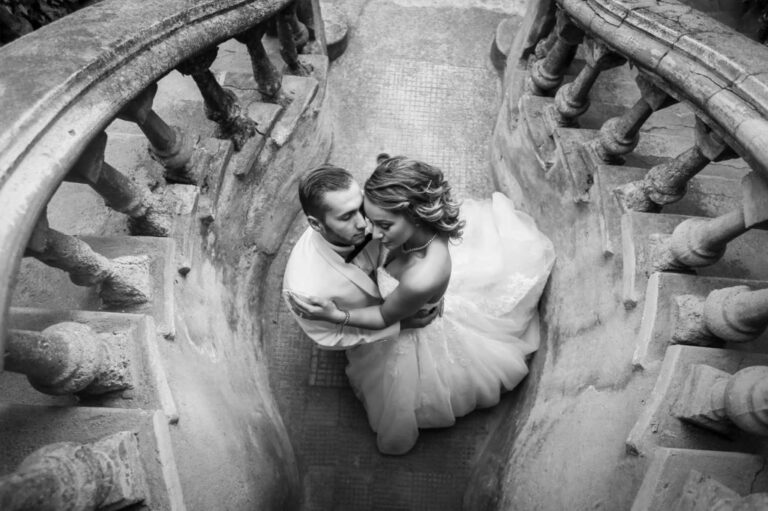 Elegant black and white photo of a newlywed couple sharing an intimate moment on a vintage staircase. Perfect for luxury wedding imagery emphasizing timeless romance and high-end venue aesthetics.
