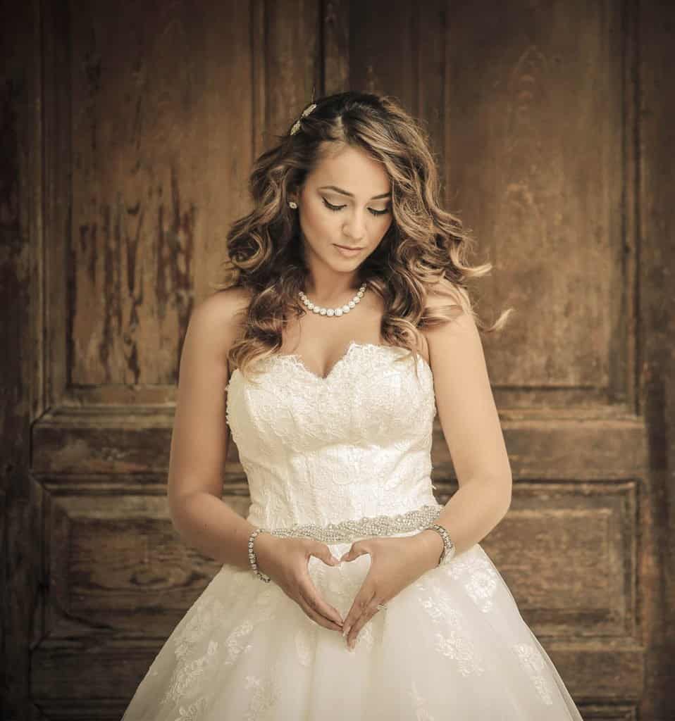 Elegant bride in a luxurious wedding gown making a heart shape with her hands against a rustic wooden background.