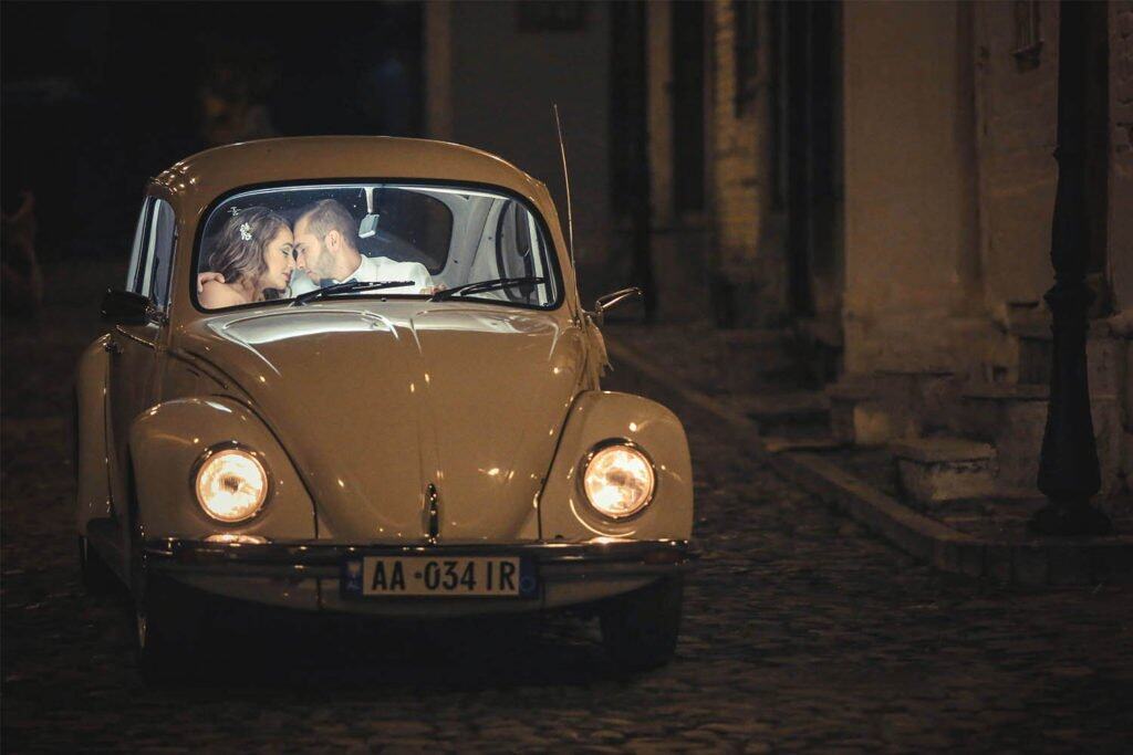 Vintage cream-colored Volkswagen Beetle with a romantic couple inside, nighttime setting, cobblestone street atmosphere for luxury wedding photography.