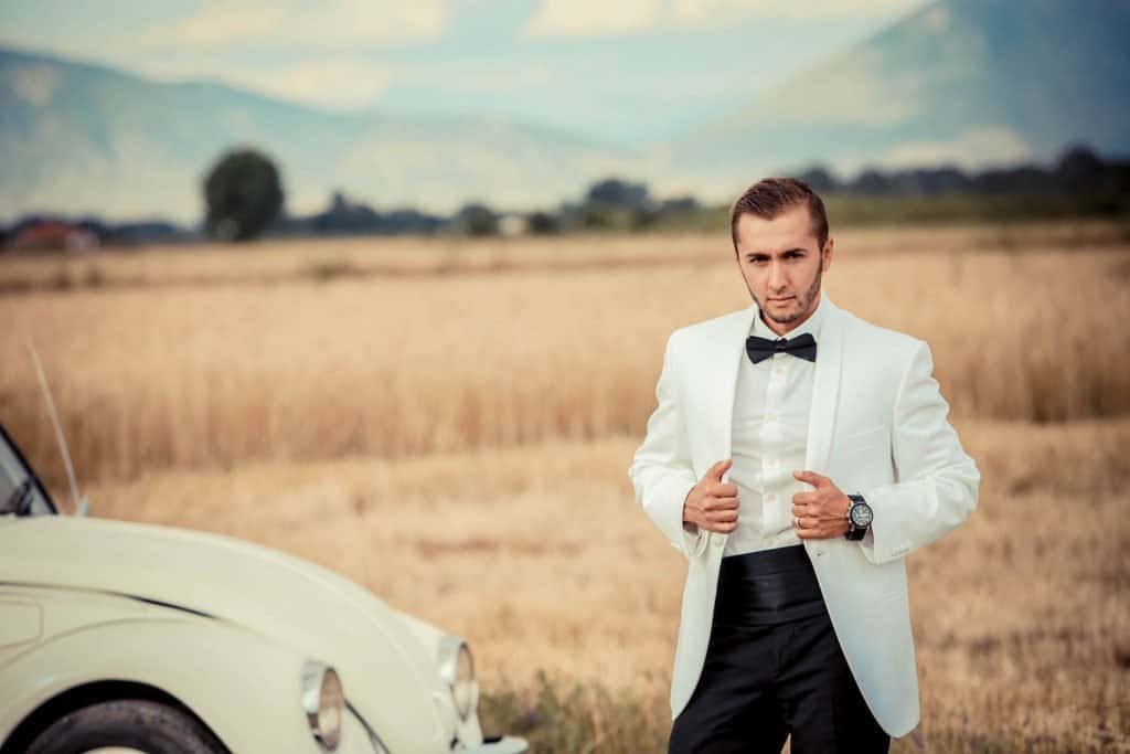 Elegant groom wearing a white tuxedo and black bow tie standing outdoors in a golden field with a vintage car, perfect for luxury wedding photos.
