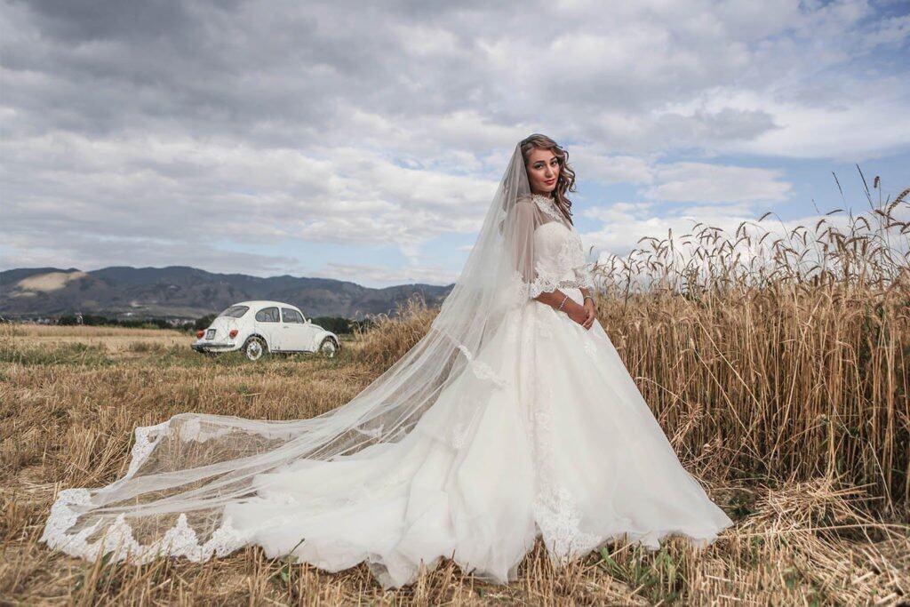 Elegant bride in a luxurious wedding gown standing in a golden field of wheat with scenic mountains in the background.