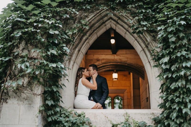Elegant wedding couple sharing romantic moment at a historic, ivy-covered stone archway in a luxurious venue.