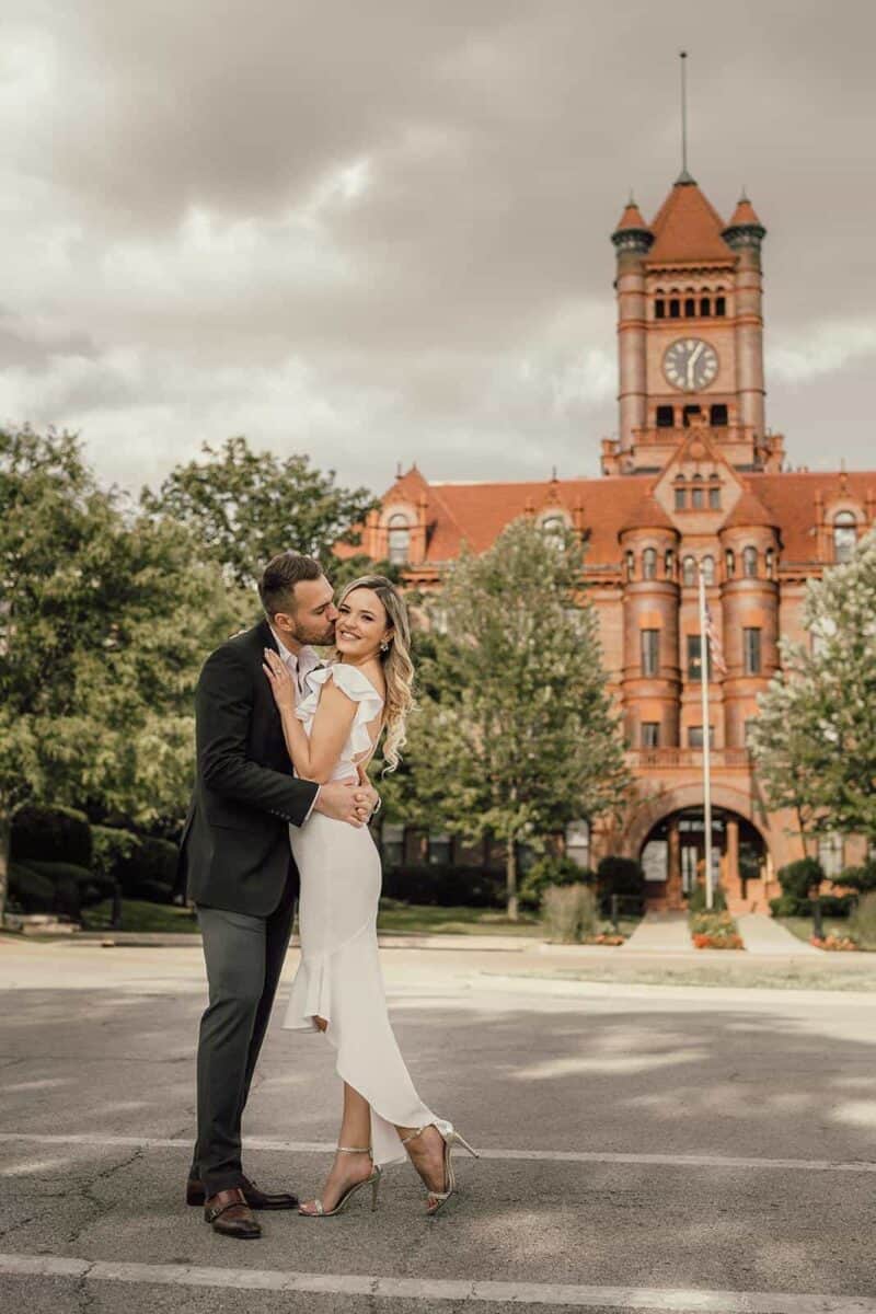 Elegant couple in wedding attire embracing in front of historic red-brick courthouse, celebrating their high-end wedding day outdoors.