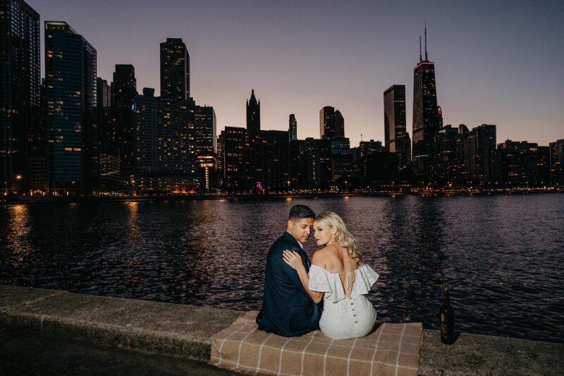 Elegant couple sitting by the river at dusk with city skyline backdrop, perfect for luxury wedding photoshoot.