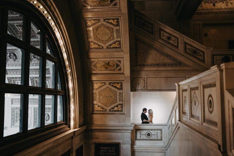 Elegant couple sharing a romantic moment on a grand staircase inside a historic upscale venue with intricate architectural details and stained glass windows.
