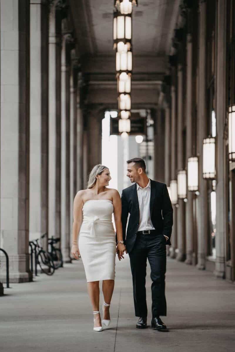 Elegant bride and groom walking hand-in-hand through a historic architectural venue, showcasing luxury wedding attire and high-end reception decor.
