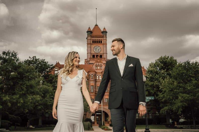 Elegant wedding couple holding hands outside historic high-end venue with clock tower, cloudy sky background.