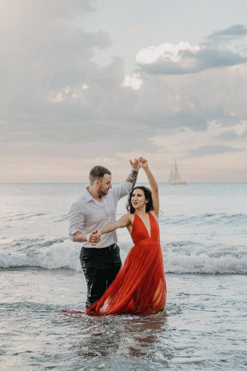 Romantic couple dancing in the ocean at sunset, dressed in elegant wedding attire, celebrating a destination wedding by the sea, high-end wedding venue, luxury beach wedding scene.