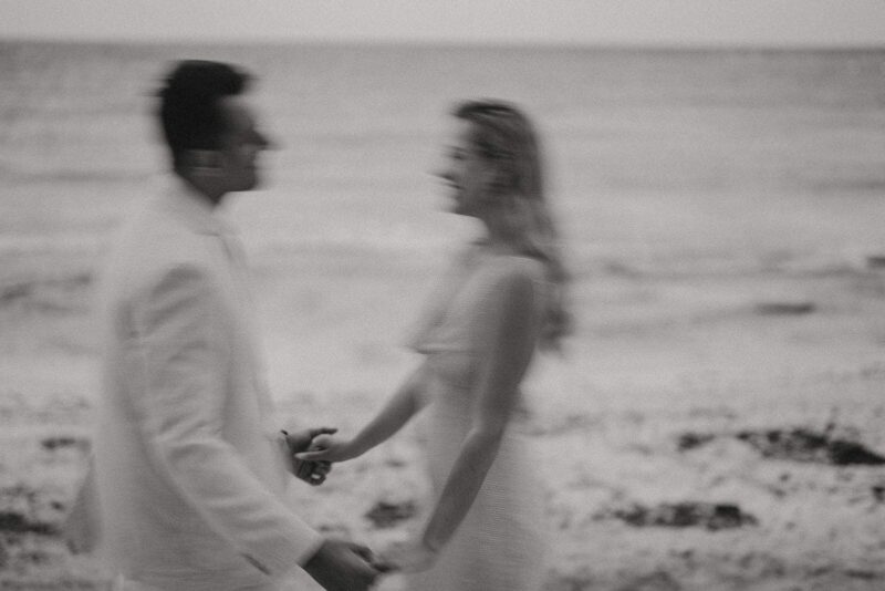 Elegant wedding couple holding hands on the beach, black-and-white portrait.