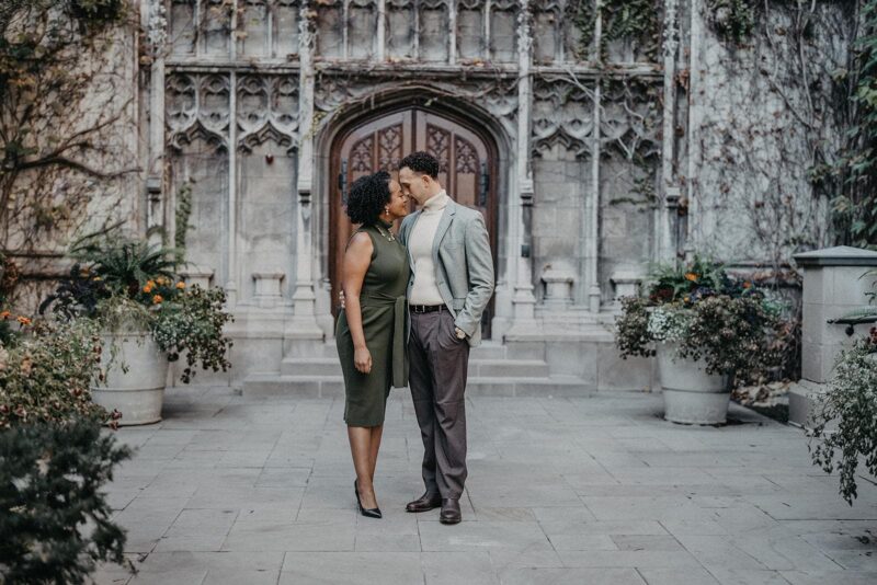 Elegant couple sharing a romantic moment outside an upscale, historic venue with gothic architectural details and lush potted greenery.