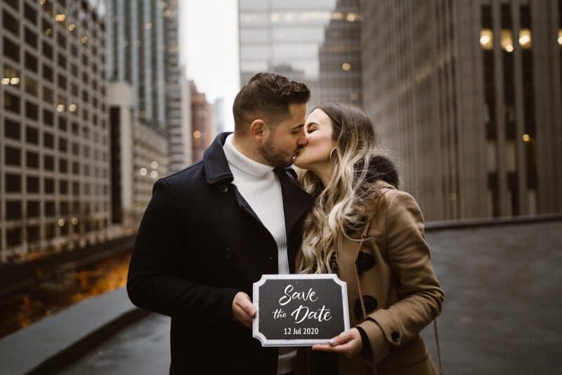 Romantic couple sharing a kiss while holding a save the date sign in a cityscape backdrop.