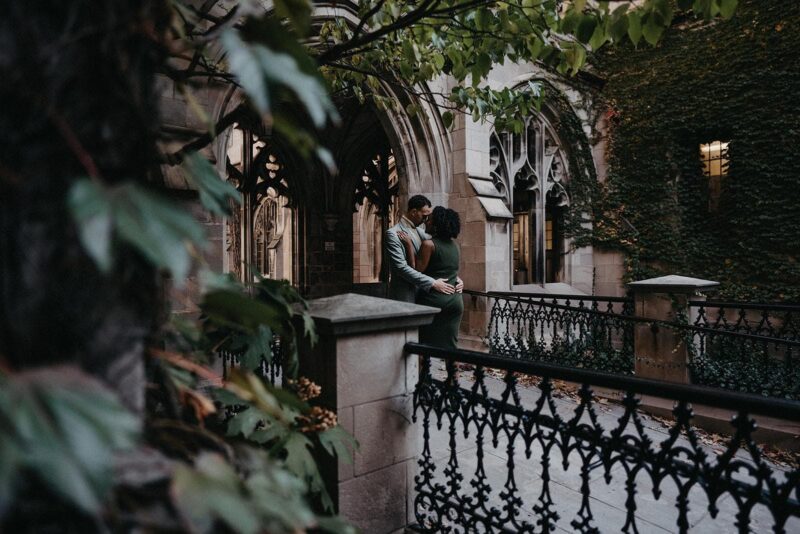 Luxurious couples embrace on a balcony of an elegant historic venue with ornate ironwork and lush greenery, perfect for upscale wedding photography.