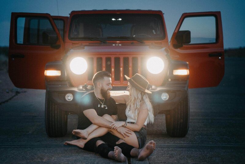 Jeep with open doors at sunset, couple enjoying romantic moment on desert road, bohemian style, adventure wedding, stunning scenic backdrop, outdoor celebration, luxury travel, high-end venue, intimate moment.