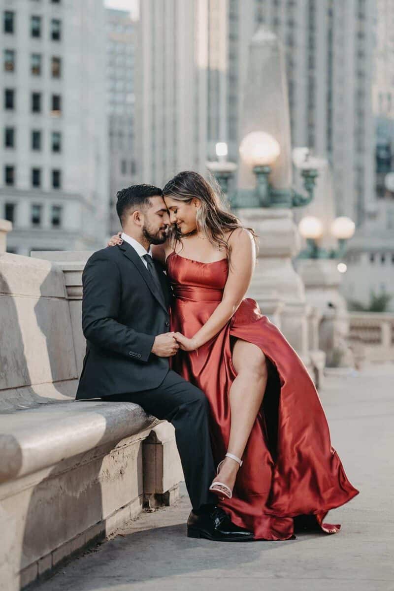Romantic couple in elegant attire sitting on city bench at dusk for luxury wedding photoshoot.
